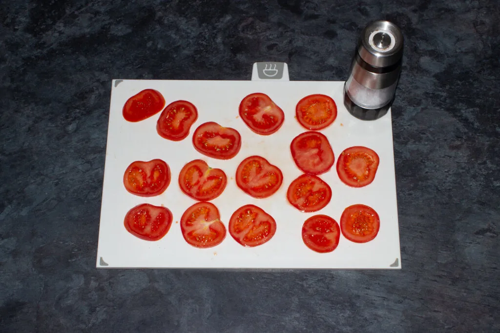 Tomato slices being sprinkled with salt on a white chopping board.