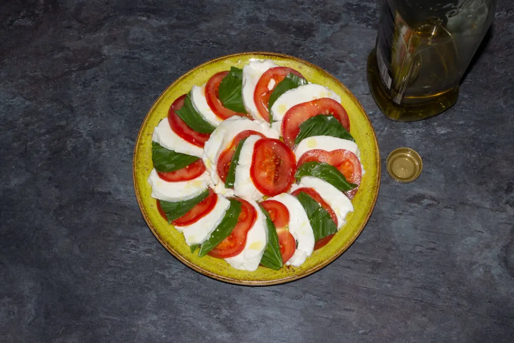 Slices of tomato, basil and mozzarella in a layered circle on a green plate with olive oil drizzled over the top.