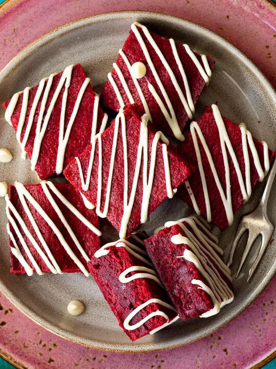Over head view of red velvet brownies drizzled with white chocolate on a cream plate set over a larger pink plate. Two brownies are on their side and there is a fork, red linen napkin and white chocolate chips in the background. Set over a green wooden backdrop.