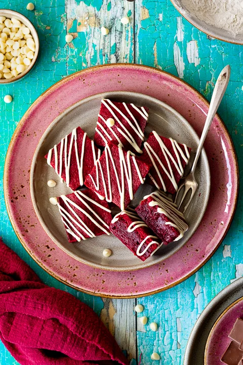 Over head view of red velvet brownies drizzled with white chocolate on a cream plate set over a larger pink plate. Two brownies are on their side and there is a fork, red linen napkin and white chocolate chips in the background. Set over a green wooden backdrop.