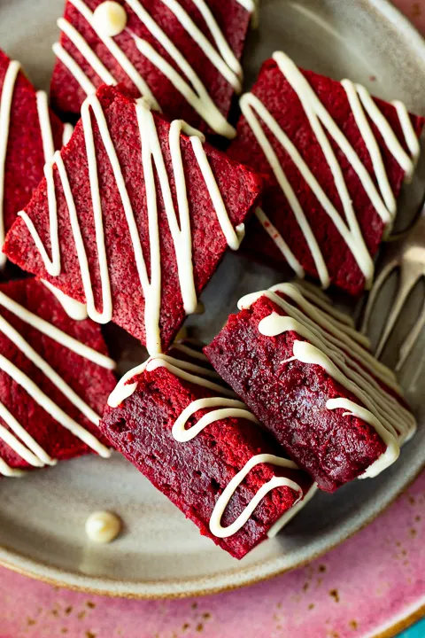 Over head close up view of red velvet brownies drizzled in white chocolate on a plate with two on their side and a fork in the background.