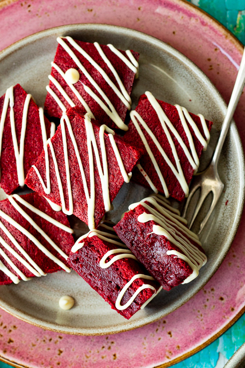 Over head view of red velvet brownies drizzled in white chocolate on a plate with two on their side and a fork in the background.