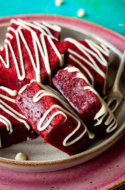 Side view of red velvet brownies drizzled in white chocolate on a plate with two on their side and a fork in the background.