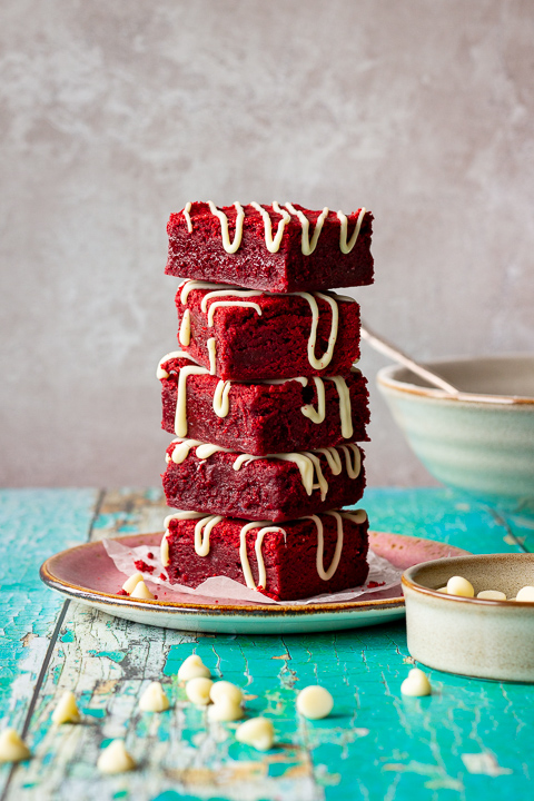 Red velvet brownies drizzled with white chocolate in a stack of 5 on a pink plate. Set over a green wooden backdrop with a bowl of flour, a bowl of white chocolate chips and scattered white chocolate chips in the background.