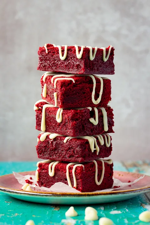 Red velvet brownies drizzled with white chocolate in a stack of 5 on a pink plate. Set over a green wooden backdrop with scattered white chocolate chips in the background.
