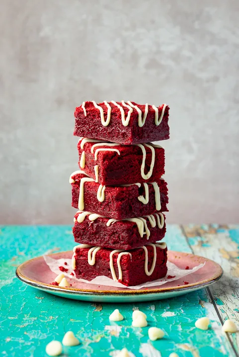 Red velvet brownies drizzled with white chocolate in a stack of 5 on a pink plate. Set over a green wooden backdrop with scattered white chocolate chips in the background.