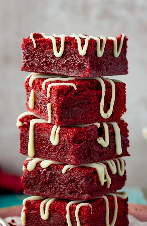Red velvet brownies drizzled with white chocolate in a stack of 5 against a light neutral backdrop.
