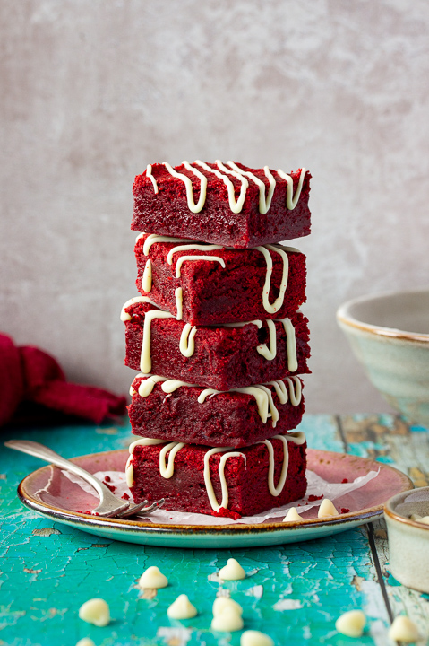 Red velvet brownies drizzled with white chocolate in a stack of 5 on a pink plate with a fork on the side. Set over a Green wooden backdrop with a red linen napkin and scattered white chocolate chips in the background.