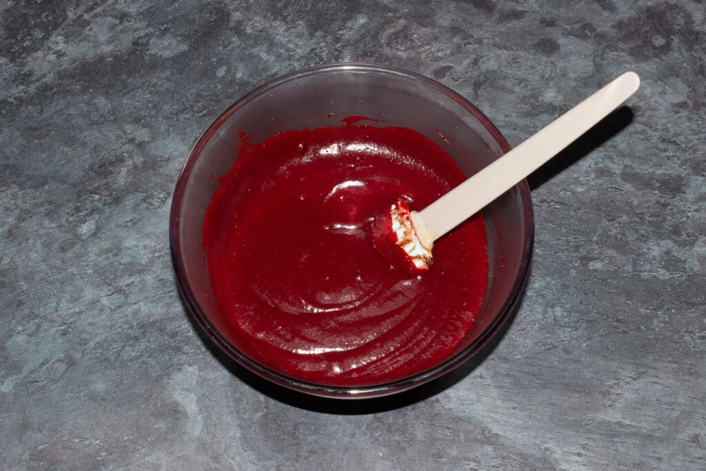 Melted butter, sugar, cocoa powder, eggs, vanilla and red food colouring mixed together in a glass bowl with a spatula.