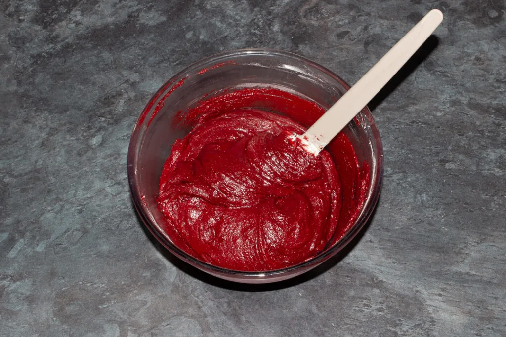 Melted butter, sugar, cocoa powder, eggs, vanilla, red food colouring, flour and salt imxed together in a glass bowl with a spatula.