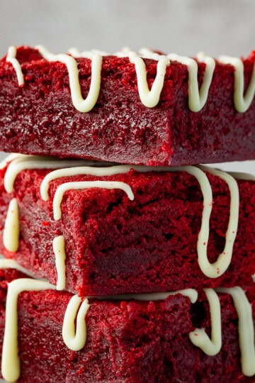Red velvet brownies drizzled with white chocolate in a stack of 3 against a light neutral backdrop.