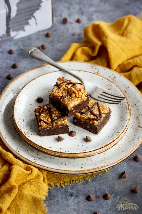 4 peanut butter brownies in a stack on top of 3 stacked plates with a fork. Set on a yellow linen napkin over a grey background, there's also scattered chocolate chips and a Bad Brownie box in the background.