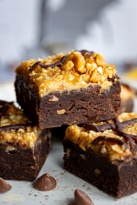 Close up of 4 peanut butter brownies on a plate. Set against a grey backdrop, there's also a Bad Brownie box, a yellow linen napkin and chocolate chips in the background.