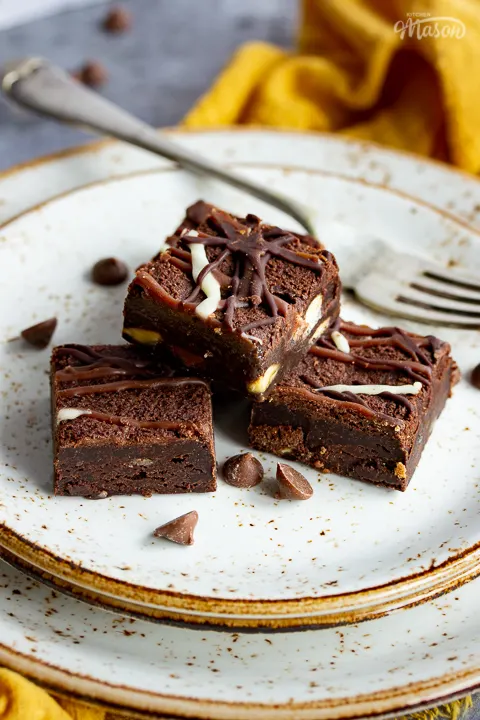 Close up of 4 triple chocolate brownies and a fork on top of 3 stacked plates. Set against a grey backdrop, there's also a Bad Brownie box, a yellow linen napkin and chocolate chips in the background.