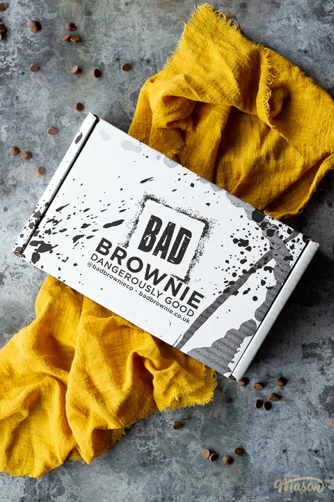 A closed box of Bad Brownie brownies on a yellow linen napkin against a grey backdrop with chocolate chips scattered around.