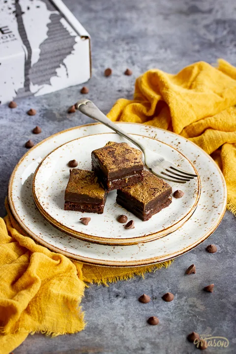 4 salted caramel brownies in a stack on top of 3 stacked plates with a fork. Set on a yellow linen napkin over a grey background, there's also scattered chocolate chips and a Bad Brownie box in the background.