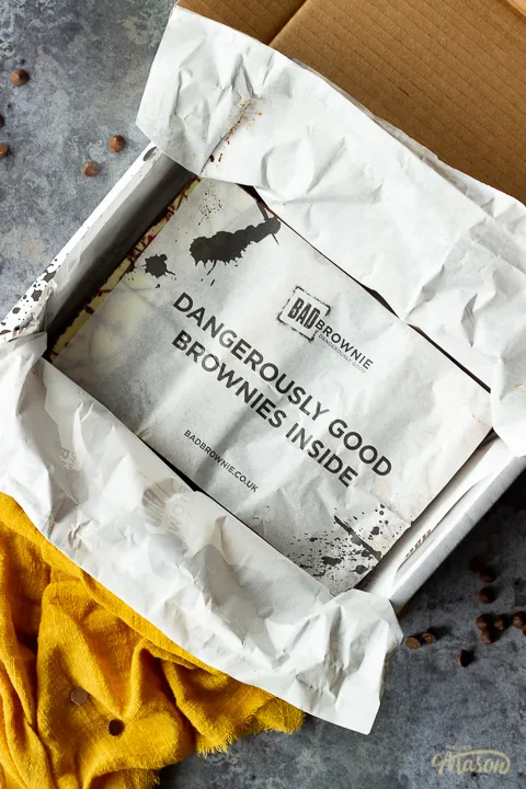 An open box of Bad Brownie brownies with a sheet saying "dangerously good brownies inside" on a yellow linen napkin against a grey backdrop with chocolate chips scattered around.