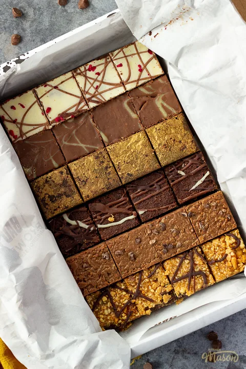 An open box of Bad Brownie brownies revealing all 6 flavours on a yellow linen napkin against a grey backdrop with chocolate chips scattered around.