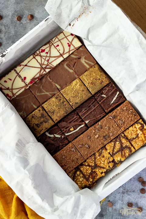 An open box of Bad Brownie brownies revealing all 6 flavours on a yellow linen napkin against a grey backdrop with chocolate chips scattered around.