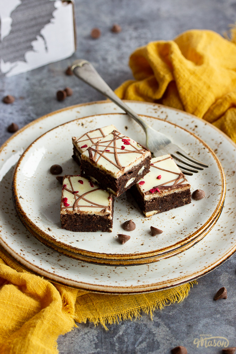 4 white chocolate raspberry brownies in a stack on top of 3 stacked plates with a fork. Set on a yellow linen napkin over a grey background, there's also scattered chocolate chips and a Bad Brownie box in the background.