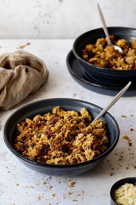 2 blue bowls containing homemade granola and spoons set over light backdrop with granola crumbs scattered everywhere. There is also a small pot with flaked almonds and a light brown napkin in the background.