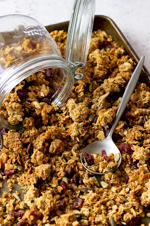 A large baking tray filled with homemade granola set over a light background. There's a glass Kilner jar and a spoon on top.