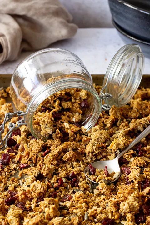 A large baking tray filled with homemade granola set over a light background. There's a glass Kilner jar and a spoon on top plus a light brown napkin and 2 stacked blue bowls in the background.