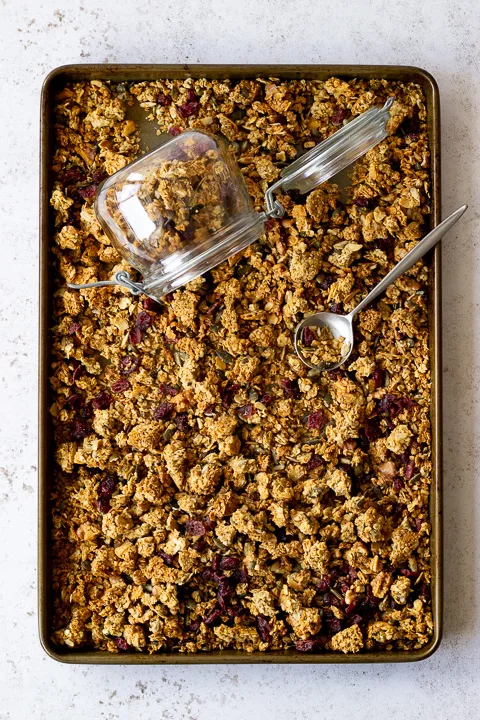 Flat lay view of a tray filled with homemade granola set over a light backdrop. There's also a glass Kilner jar and a spoon in the granola itself.