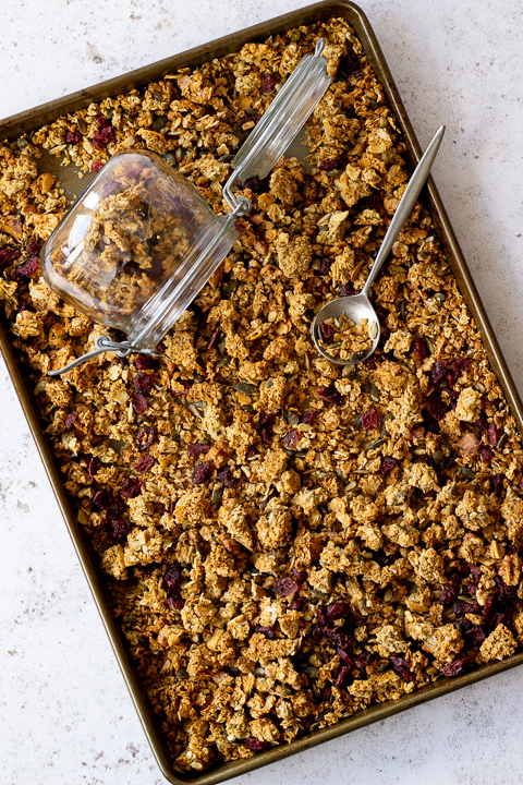 Angled flat lay view of a tray filled with homemade granola set over a light backdrop. There's also a glass Kilner jar and a spoon in the granola itself.