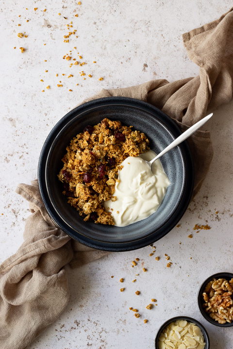 A flat lay view of a blue bowl containing homemade granola, yoghurt and a spoon. The bowl is set on a blue plate on a light brown napkin over a light backdrop. There are granola crumbs scattered everywhere and two small pots with walnuts and almonds in them.
