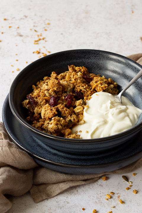 Close up side view of a blue bowl filled with homemade granola, yoghurt and a spoon set on a blue plate over a light brown napkin. It's set on a light backdrop with granola crumbs scattered around.