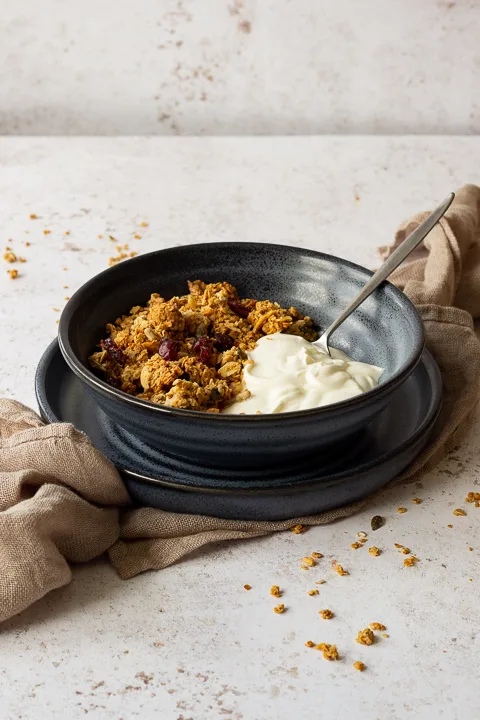 Side view of a blue bowl filled with homemade granola, yoghurt and a spoon set on a blue plate over a light brown napkin. It's set on a light backdrop with granola crumbs scattered around.