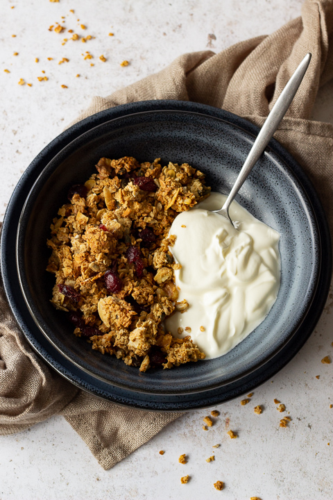 A close up flat lay view of a blue bowl containing homemade granola, yoghurt and a spoon. The bowl is set on a blue plate on a light brown napkin over a light backdrop. There are granola crumbs scattered everywhere.