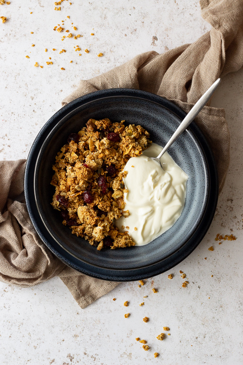 A flat lay view of a blue bowl containing homemade granola, yoghurt and a spoon. The bowl is set on a blue plate on a light brown napkin over a light backdrop. There are granola crumbs scattered everywhere.