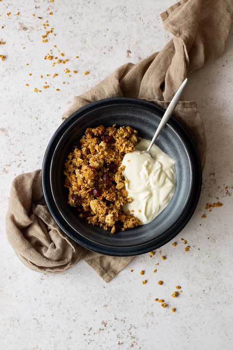 A distanced flat lay view of a blue bowl containing homemade granola, yoghurt and a spoon. The bowl is set on a blue plate on a light brown napkin over a light backdrop. There are granola crumbs scattered everywhere.