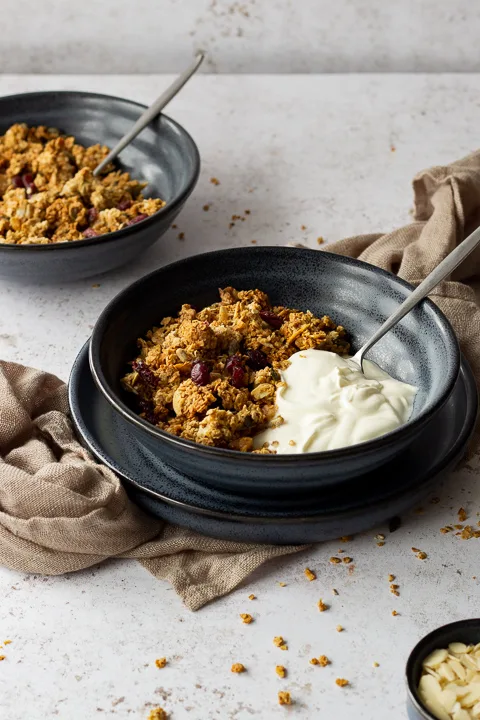 Side view of a blue bowl filled with homemade granola, yoghurt and a spoon set on a blue plate over a light brown napkin. It's set on a light backdrop with another blue bowl filled with granola, a small pot filled with flaked almonds and granola crumbs scattered around.