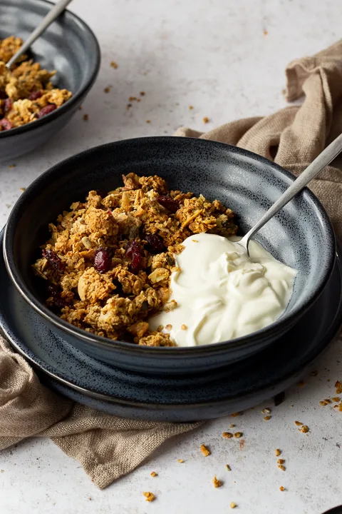 Side view of a blue bowl filled with homemade granola, yoghurt and a spoon set on a blue plate over a light brown napkin. It's set on a light backdrop with another blue bowl filled with granola and granola crumbs scattered in the background.