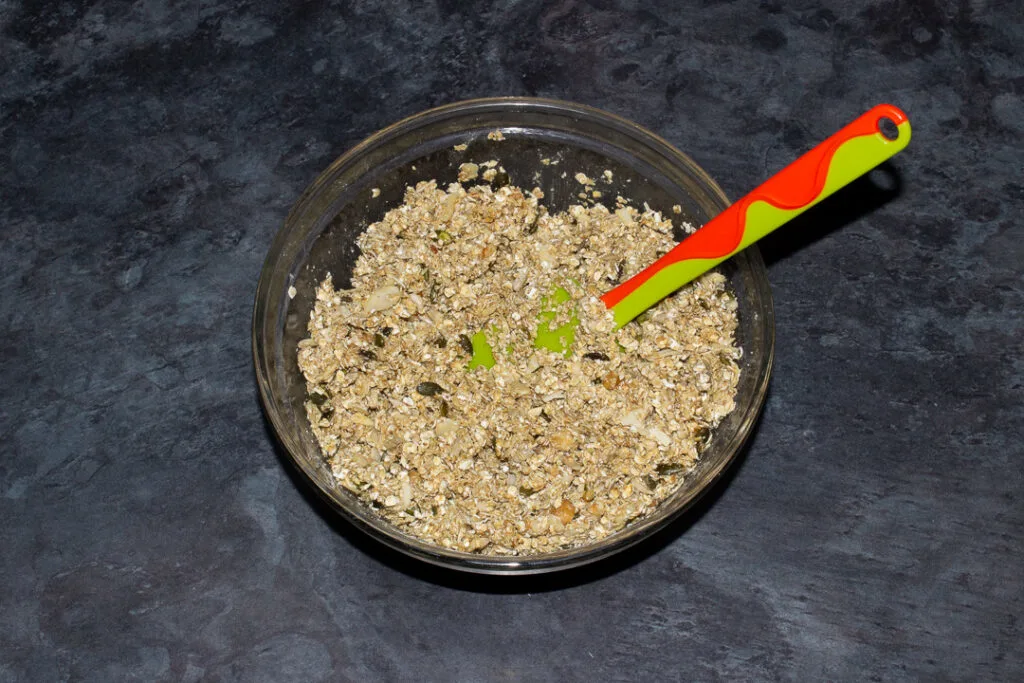 A glass bowl with homemade granola ingredients and a spatula inside.