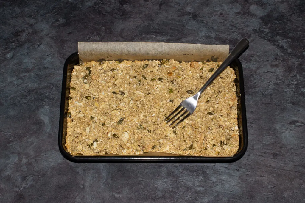 A lined rectangular tin with part baked homemade granola in it and a fork resting on top