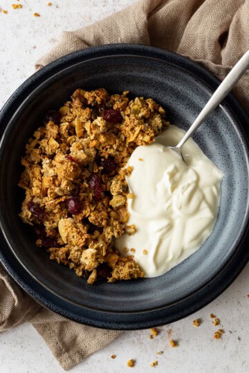 A flat lay view of a blue bowl containing homemade granola, yoghurt and a spoon. The bowl is set on a blue plate on a light brown napkin over a light backdrop. There are granola crumbs scattered everywhere.