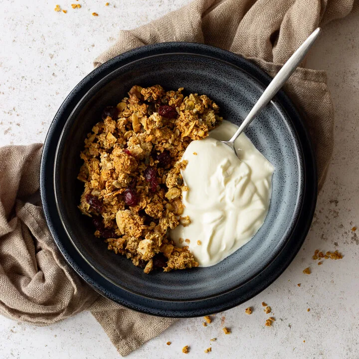 A flat lay view of a blue bowl containing homemade granola, yoghurt and a spoon. The bowl is set on a blue plate on a light brown napkin over a light backdrop. There are granola crumbs scattered everywhere.