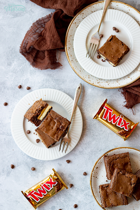 Flat lay view of 3 Twix brownies and a fork stacked on a white ribbed plate over a mottled grey backdrop. There are 2 Twix bars, a brown linen napkin, two other plates of Twix brownies and chocolate chips scattered in the background.