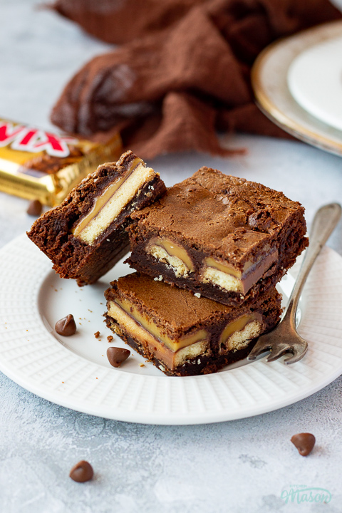 3 Twix brownies and a fork stacked on a white ribbed plate over a mottled grey backdrop. There is a Twix bar, a brown linen napkin, another plate of Twix brownies and chocolate chips scattered in the background.