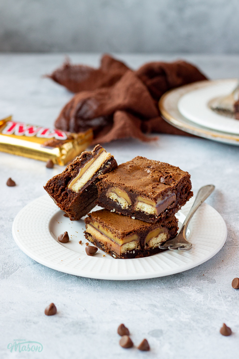 3 Twix brownies and a fork stacked on a white ribbed plate over a mottled grey backdrop. There is a Twix bar, a brown linen napkin, another plate of Twix brownies and chocolate chips scattered in the background.