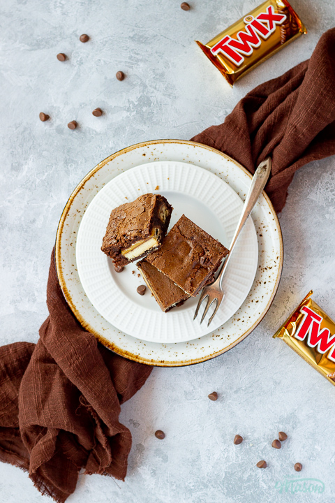 Flat lay view of 3 Twix brownies and a fork stacked on a white ribbed plate over a mottled grey backdrop. There are 2 Twix bars, a brown linen napkin and chocolate chips scattered in the background.