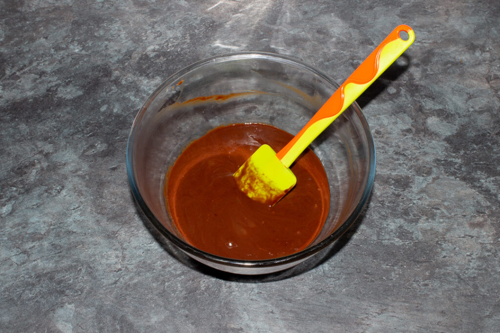Melted butter, dark chocolate and eggs mixed together in a glass bowl with a green spatula.