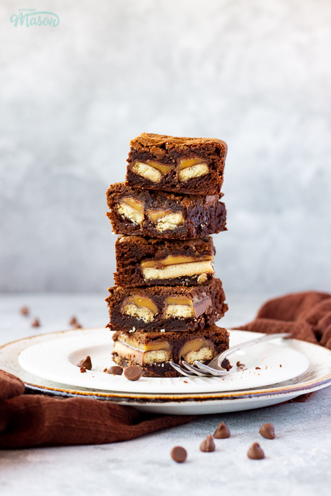 A stack of 5 Twix brownies on a white ribbed plate with a fork set over a grey mottled backdrop. There's a brown linen napkin and chocolate chips scattered in the background.
