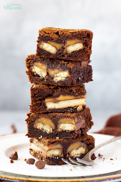A stack of 5 Twix brownies on a white ribbed plate with a fork set over a grey mottled backdrop. There's a brown linen napkin and chocolate chips scattered in the background.