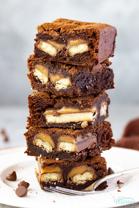 Close up of a stack of 5 Twix brownies on a white ribbed plate with a fork set over a grey mottled backdrop. There's a brown linen napkin and chocolate chips scattered in the background.