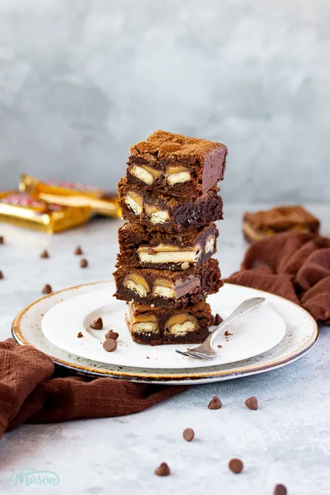 A stack of 5 Twix brownies on a white ribbed plate with a fork set over a grey mottled backdrop. There's a brown linen napkin, a Twix bar and chocolate chips scattered in the background.